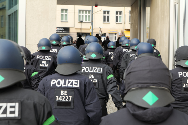 Eine Gruppe von Polizisten in Uniform und Helmen steht vor einem Gebäude mit einigen Menschen, Pfählen und anderen Gebäuden im Hintergrund.