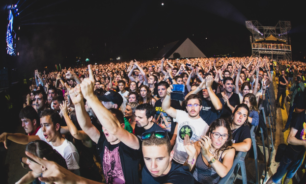 Menschenmenge auf einem Musikfestival mit erhobenen Händen, sitzend und stehend, einem Metallzaun auf der rechten Seite, Gebäuden und Lichtern im Hintergrund und einem dunklen Himmel.