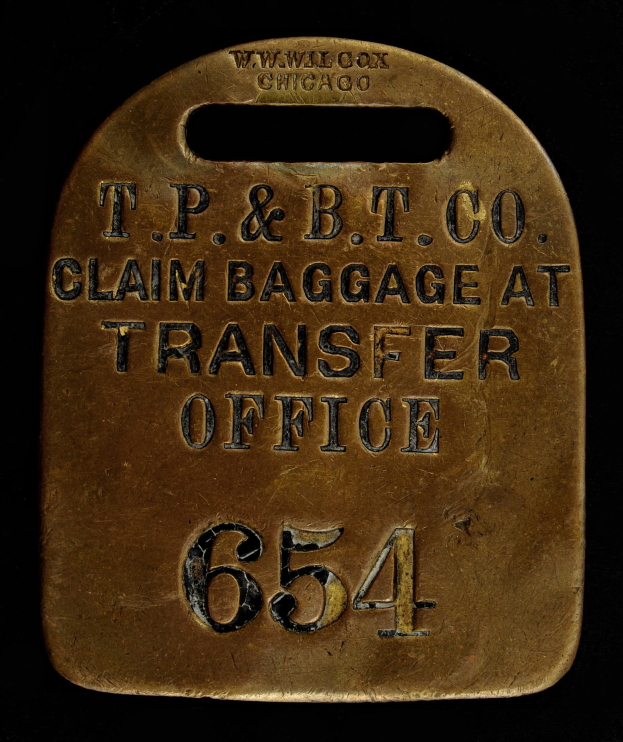 Brass luggage tag with "T.P. & B.T. Co. Claim Baggage at Transfer Office" engraved, set against a dark background.