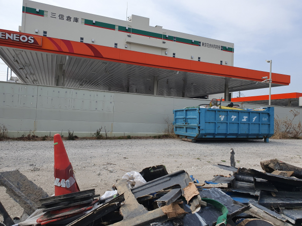 Großes Gebäude mit Bergen von Schrott, einem Verkehrskegel und einem blauen Container davor, umgeben von Pflanzen und Pfählen während der Abrissarbeiten.