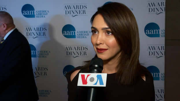 Emmy Rossum, eine Frau in einem schwarzen Kleid, hält ein Mikrofon mit einem Mann zu ihrer Linken und einem Banner mit der Aufschrift 'Aam Awards Dinner' im Hintergrund.