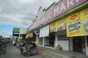 Eine belebte Stadtstraße mit parkenden Fahrzeugen, Fußgängern, Gebäuden, Strommasten, Bäumen und einem bewölkten Himmel, mit einem Geschäft mit einem Schild, auf dem "Bongabon Public Market" steht.