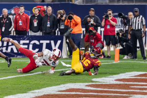 Ein Fussballspieler taucht in die Endzone für einen Touchdown, mit einer Gruppe von Menschen am Boden, einige halten Kameras, und ein Banner mit Text im Vordergrund, während Geländer, Pfosten und andere Gegenstände im Hintergrund sichtbar sind.