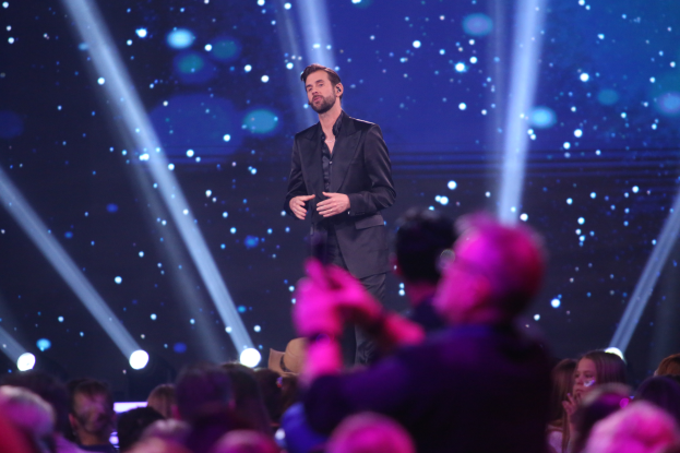 Ein Mann in einem schwarzen Anzug steht auf einer hell erleuchteten Bühne und wird als Gewinner von 'Dancing with the Stars' identifiziert.