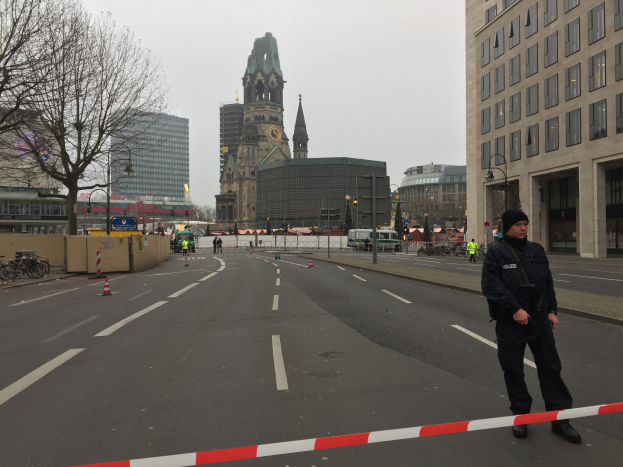 Ein Polizist in schwarzer Uniform und Mütze steht in einer Berliner Straße umgeben von Absperrbaken, Fahrrädern, Laternenpfählen, Bäumen und Gebäuden, mit einem Uhrenturm und einem klaren blauen Himmel im Hintergrund.