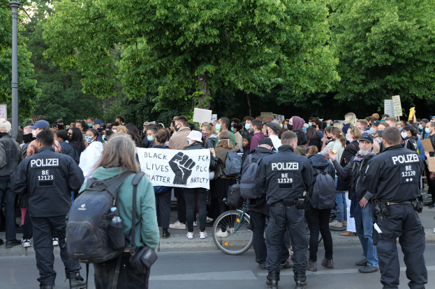 Eine große Gruppe von Menschen steht auf der Straße, einige halten Schilder und andere tragen Mützen und Taschen, mit einem Fahrrad im Vordergrund und Bäumen und einem Pfahl im Hintergrund.