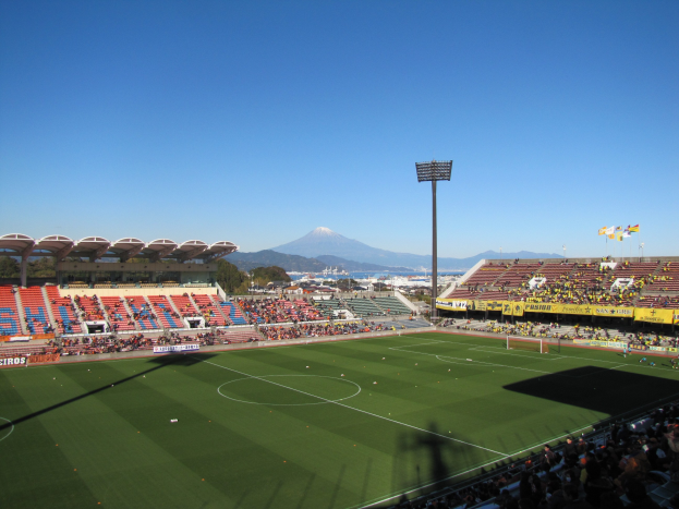 Ein hell erleuchtetes Fußballstadion mit Zuschauern auf den Tribünen, umgeben von Bäumen und Hügeln, unter einem sichtbaren Himmel, mit Laternenmasten, Bannern und Fahnen.