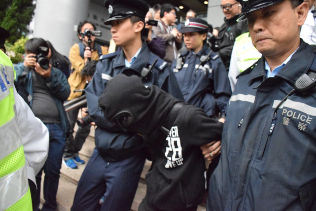 Eine Gruppe von Polizeibeamten vor einer Menschenmenge vor einem Gebäude mit Säulen und Deckenbeleuchtung, mit Bäumen links im Bild.