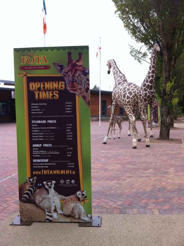 Giraffen vor B├Ąumen, H├╝tten und Flaggen mit einer Tafel mit Text, Logos und Tierbildern.