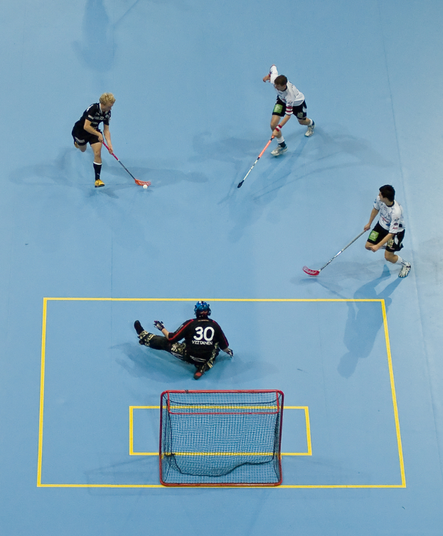 Drei Personen in Sportkleidung spielen Hockey, wobei eine Person rechts mit einem Hockeystock einen Ball schießt und eine Person hinter einem Netz auf dem Boden sitzt.