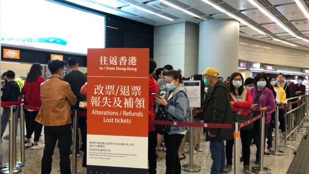 Eine Gruppe von Menschen in Masken steht in einer Schlange an einem Flughafen, umgeben von Absperrpollern mit Bändern, mit einer Tafel im Vordergrund, auf der "Hong Kong Airport Refunds Due to Covid-19" steht, und Säulen und Deckenleuchten im Hintergrund.
