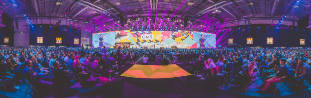 Eine große Menschenmenge sitzt auf Stühlen vor einer hell erleuchteten Bühne mit mehreren stehenden Personen, wahrscheinlich bei einem Online-Marketing-Rockstars-Event.