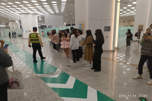 Menschen in einer Reihe an einem Flughafen-Check-in-Schalter, einige halten Taschen und Mobiltelefone, mit Säulen und Deckenleuchten im Hintergrund.