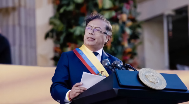 Ein Mann in einem blauen Anzug und Krawatte steht an einem Rednerpult, hält Papiere und spricht in ein Mikrofon, mit einer Pflanze und einer Wand dahinter, was eine formelle Umgebung suggeriert.