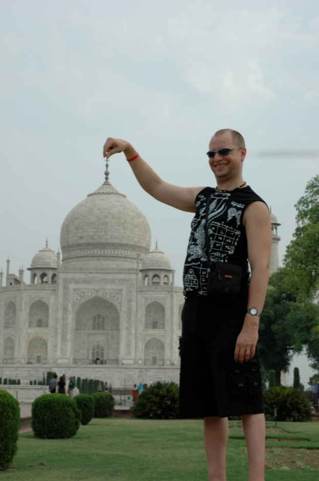 Eine Person mit Brille steht und lächelt vor dem Taj Mahal, mit Menschen, Pflanzen, Bäumen, Boden und Himmel im Hintergrund.