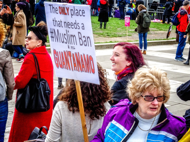 Eine Gruppe von Menschen steht auf dem Boden, einige halten Taschen, während eine Frau ein Schild hält, auf dem steht: "Der einzige Ort, an dem ein Muslimverbot benötigt wird, ist Guantanamo", vor einem Metallzaun, Gras, einem Schild, Pfosten, einem Gebäude mit Fenstern und Bäumen.