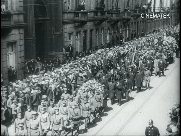 Schwarzes Foto einer Parade mit einer großen Menge, die eine Straße entlangmarschiert, einige halten Gewehre, vor einem Gebäude mit einem sichtbaren Wasserzeichen.