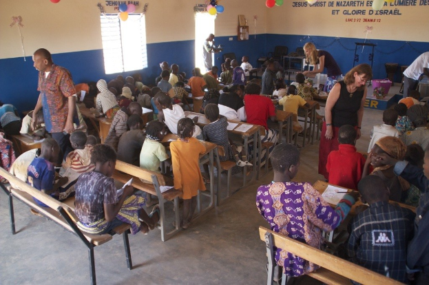 Kinder auf Bänken in der Nähe von Erwachsenen, während Stühle, eine Tür, ein Fenster, ein Tisch, Luftballons und Text im Hintergrund zu sehen sind.