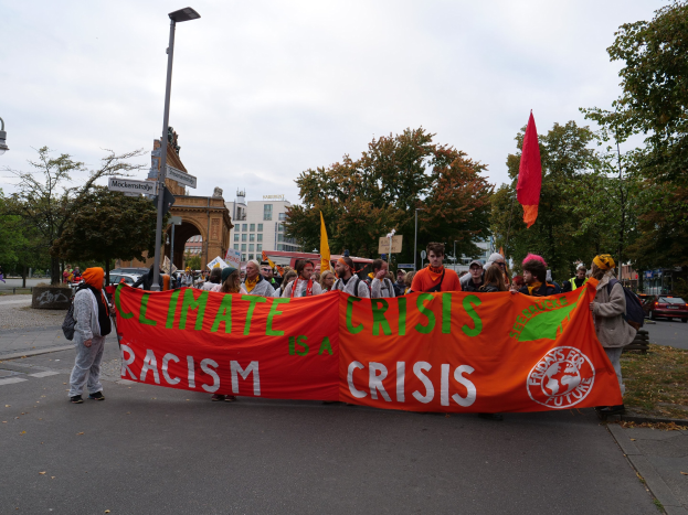 Eine Gruppe von Menschen marschiert auf einer von Bäumen gesäumten Straße, die ein Banner mit der Aufschrift 'Klimakrise ist eine Krise' trägt, mit vorbeifahrenden Fahrzeugen und Gebäuden im Hintergrund unter einem klaren blauen Himmel.