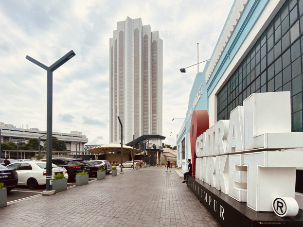 Ein belebter zentraler Markt in Kuala Lumpur, Malaysia, mit hohen Gebäuden, Straßenlaternen, fahrenden Fahrzeugen, Fußgängern auf dem Gehweg, sichtbaren Namensschildern, Bäumen und einem bewölkten Himmel.