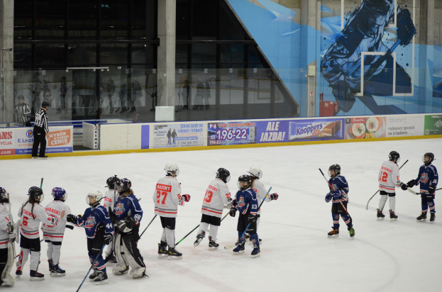 Gruppe junger Menschen beim Eishockey auf einer Indoor-Eisfläche, mit Helmen und Stöcken, mit einem Zuschauer links und Bannern, Säulen und Glaswänden im Hintergrund.