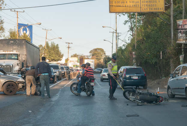 Gruppe von Menschen, die um ein verunglücktes Motorrad auf der Seite der Stra├če stehen, mit vorbeifahrenden Fahrzeugen, B├Ąumen, Masten und einem klaren Himmel im Hintergrund.