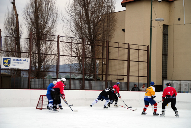 Menschen beim Eisspiel auf einer Eisbahn mit Gebäuden, Bäumen, einer Straßenlaterne, einem Namensschild und Zäunen im Hintergrund unter einem Himmel.