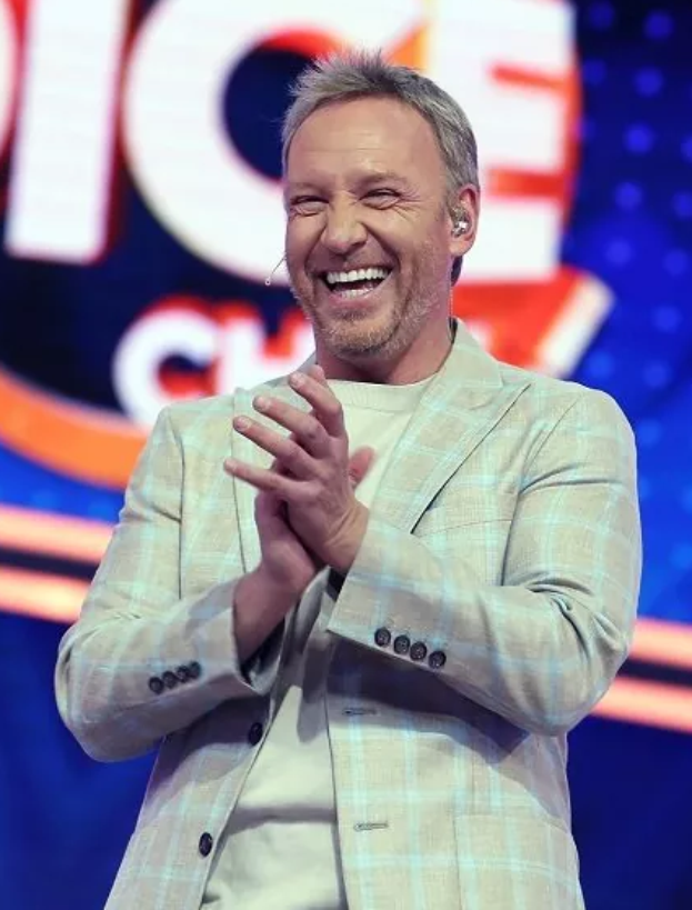 Ein Mann in einem Anzug steht auf einer Bühne, lächelt und klatscht, mit einem Schild im Hintergrund, auf dem "The Voice" steht.