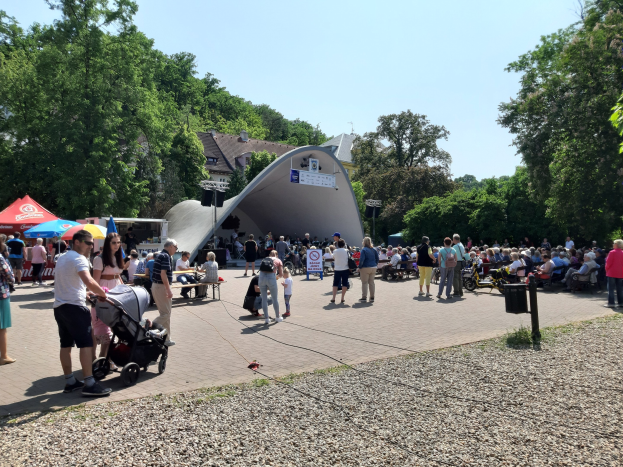 Eine Menschenmenge versammelt sich vor einem Gebäude bei einer Festivität, einige sitzen auf Bänken, andere stehen; ein Kinderwagen ist im Vordergrund, und Bäume, Gebäude und ein klarer blauer Himmel sind im Hintergrund zu sehen.