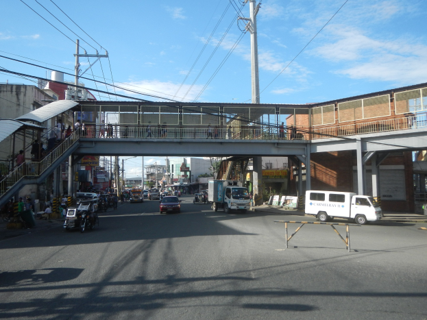 Stadtstraße mit fahrenden Fahrzeugen, eine Fußgängerbrücke mit Menschen, Strommasten mit Drähten, Gebäude mit Fenstern und ein bewölkter Himmel.