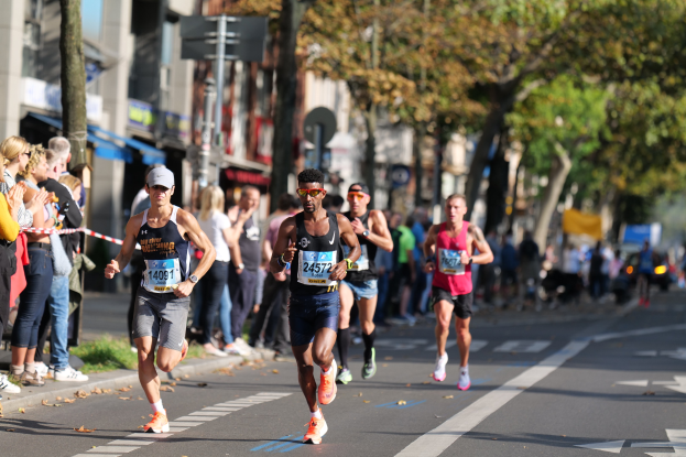 Eine Gruppe von Marathonläufern in Turnschuhen und Hüten auf einer Stadtstraße, mit Zuschauern auf der linken Seite und Bäumen, Gebäuden und einem klaren blauen Himmel im Hintergrund.