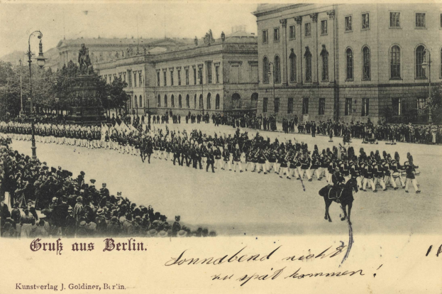 Ein altes Schwarz-Weiß-Foto eines Umzugs in Berlin, Deutschland, mit Menschen auf dem Boden und auf Pferden, einer Statue auf einem Podest, Straßenlaternen, Bäumen, Gebäuden mit Fenstern und einem bewölkten Himmel, mit Text am unteren Bildrand.