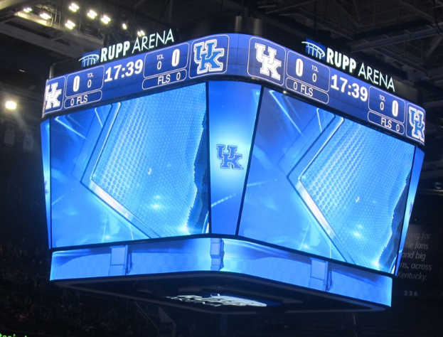 Großer Bildschirm auf einem Basketballfeld, das "Kentucky Wildcats vs Florida State Seminoles" anzeigt, mit Zuschauern auf den Tribünen und Beleuchtung von oben.