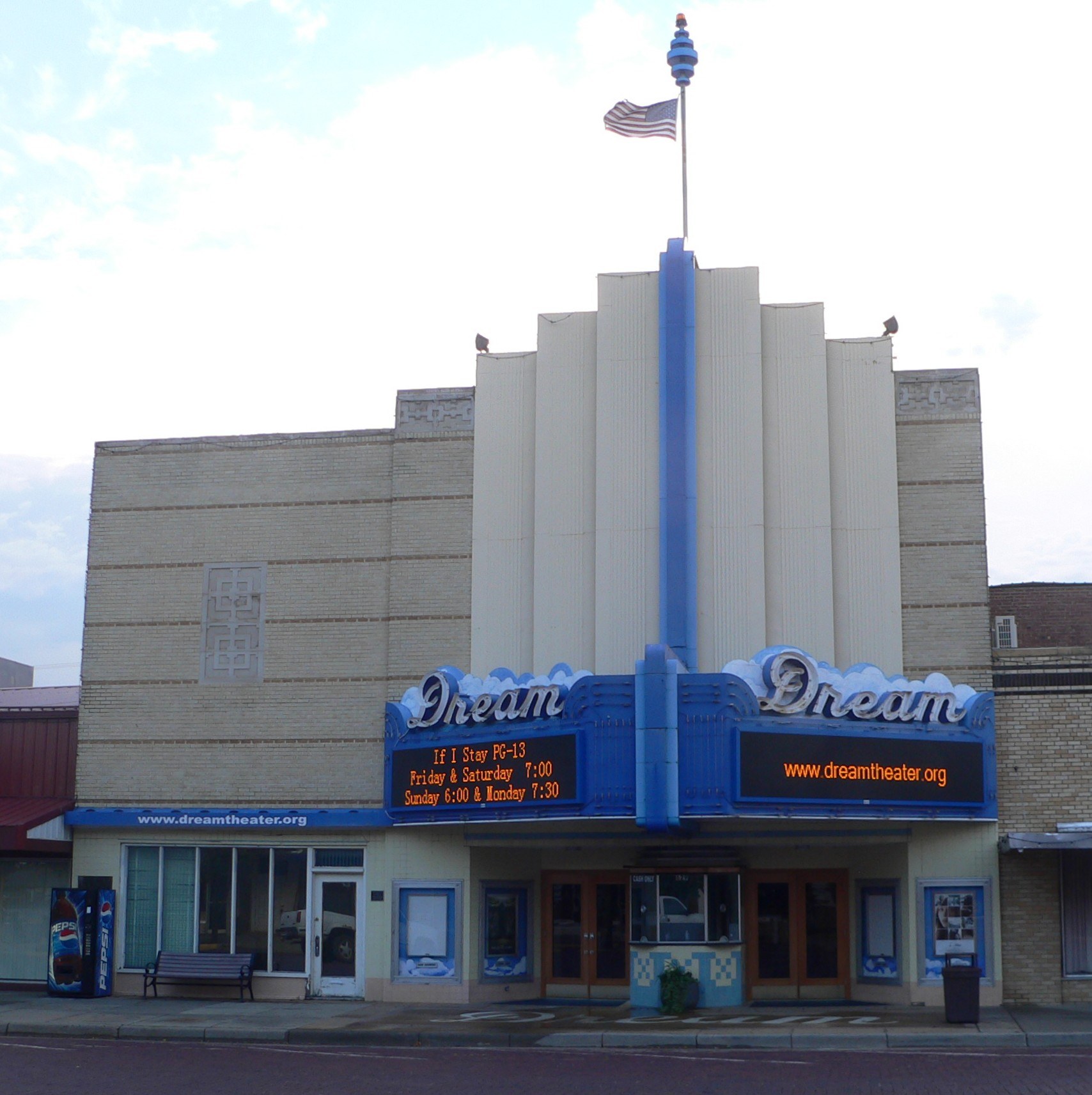 Außenansicht des Dream Theaters in St. Louis, Missouri, mit Glastüren und -fenstern, einem Namensschild, einer Bank, einem Mülleimer und einer Flagge auf dem Gebäude vor einem bewölkten Himmel.