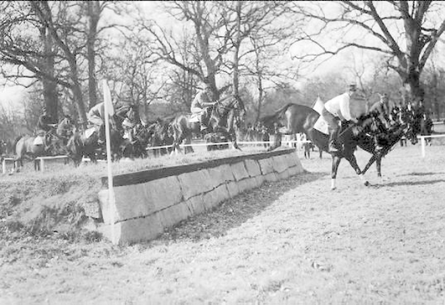Schwarzes und weißes Foto von Menschen, die auf Pferden über eine grüne Wiese reiten, mit einer Wand und Stangen im Vordergrund, Bäumen und Himmel im Hintergrund und Text unten, der "Pferdespringen bei der National Horse Show in Washington, D.C. USA" lautet.