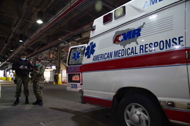 American Medical Response ambulance parked indoors with two masked individuals in front, a vehicle to the left, and additional people and objects in the background.