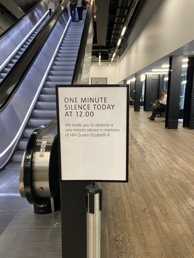Eine Rolltreppe im Flughafen mit einem Schild, auf dem "Eine Minute Stille heute" steht, einige Menschen darauf und Deckenleuchten im Hintergrund.