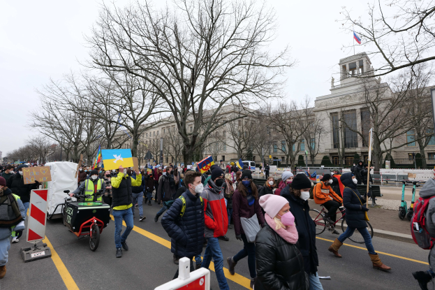 Eine große Gruppe von Menschen marschiert auf einer Straße in Washington, D.C. und hält Schilder und fährt Fahrräder, mit Bäumen und einem klaren blauen Himmel im Hintergrund.