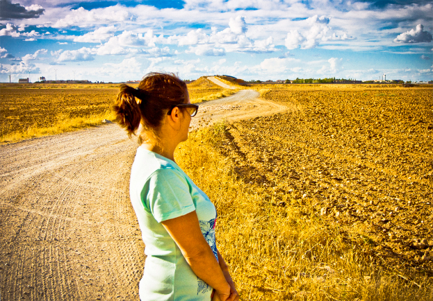 Eine Frau steht auf einem schlammigen Weg, der von landwirtschaftlichen Flächen und Bäumen auf beiden Seiten gesäumt ist.