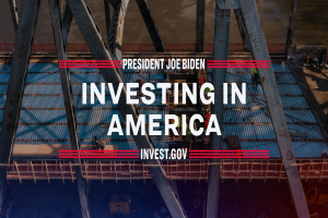 Präsident Joe Biden in der Nähe einer Brücke mit dem Text "In America investieren" im Vordergrund, umgeben von verstreuten Gegenständen.