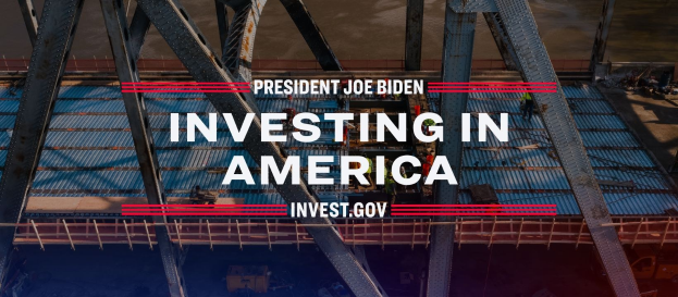 Präsident Joe Biden in der Nähe einer Brücke mit dem Text "In America investieren" im Vordergrund, umgeben von verstreuten Gegenständen.