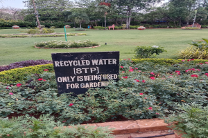 Schild mit der Aufschrift "Recycled Water (STP) Only Being Used for Gardening" in einem Garten mit grünem Gras, bunten Blumen, hohen Bäumen und einem klaren blauen Himmel.