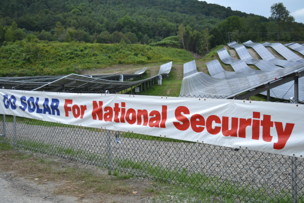 Ein Zaun mit einer 'Gehen Sie Solar für die nationale Sicherheit' -Plakette vor einem Solarpanel-Feld, Bäumen, Pflanzen und Gras unter einem klaren blauen Himmel.