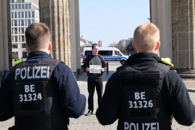 Drei Polizeibeamte stehen vor einem Gebäude, mit einem Mann, der ein Blatt Papier zwischen ihnen hält, flankiert von Säulen und vor dem Hintergrund von Fahrzeugen, Menschen, Bäumen, Gebäuden und Himmel.