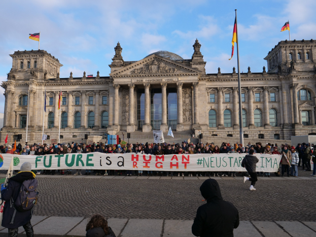 Gruppe von Menschen mit einem Banner mit der Aufschrift "Zukunft ist ein Mensch Neustar ima" vor dem Reichstaggebäude in Berlin, Deutschland, mit sichtbaren architektonischen Details und umgeben von Flaggen unter einem bewölkten Himmel.
