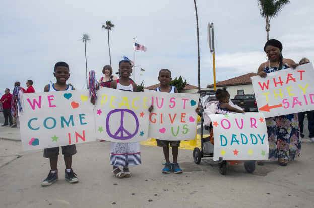 Eine Gruppe von Kindern hält Schilder in der Hand und steht vor einem Gebäude, während eine Frau einen Kinderwagen mit einem Baby schiebt; auf einem Schild steht "Wir kommen in Frieden, gebt uns unseren Daddy."