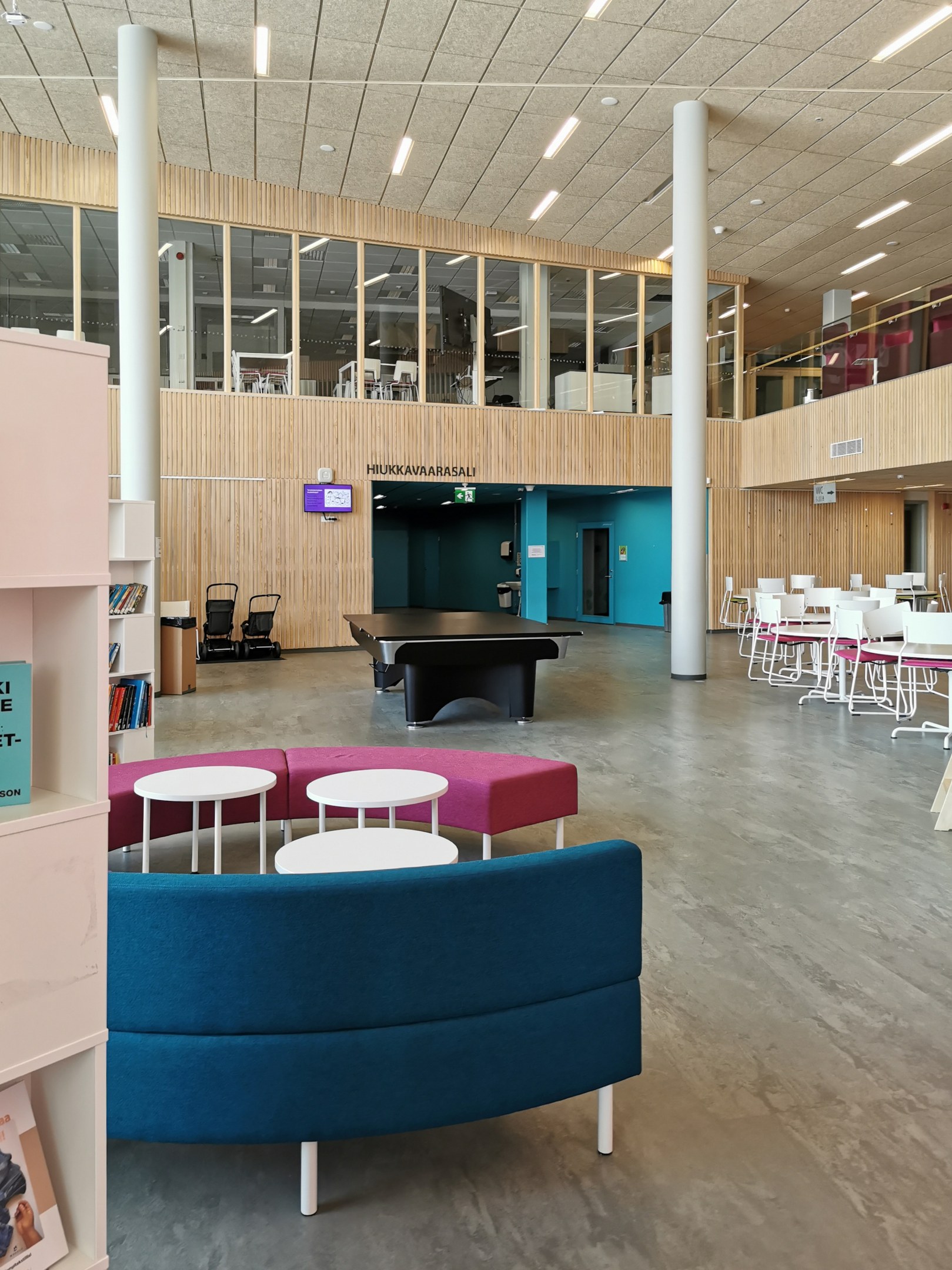 Ein geräumiger Raum, der an eine Schulbibliothek erinnert, mit Tischen, Stühlen, Sofas, Hockern, Bücherregalen, Deckenleuchten, einem Wandfernseher und Säulen.