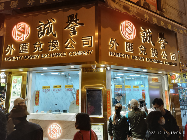 Eine Gruppe von Menschen steht vor einem hell erleuchteten Gebäude in der Nacht, mit Schildern, einige tragen Taschen und ein Wasserzeichen im rechten unteren Eck.