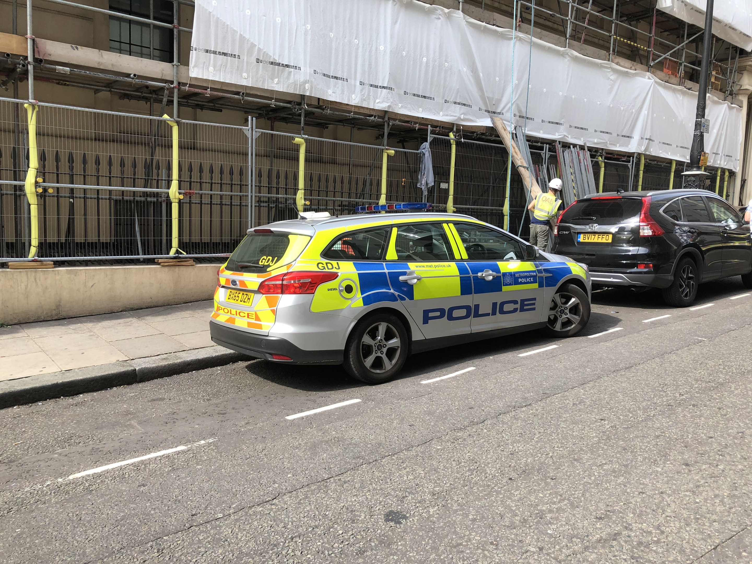 Polizeiauto auf einer Straße mit zwei Helikopterpiloten und einem weiß verhüllten Gebäude im Hintergrund, wahrscheinlich an einem Tatort in London.