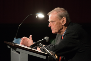 Ein Mann im Anzug und Krawatte steht an einem Rednerpult mit einem Mikrofon und einem Blatt Papier, beleuchtet von einer an der Stange befestigten Lampe, was darauf hindeutet, dass er Ökonom ist.